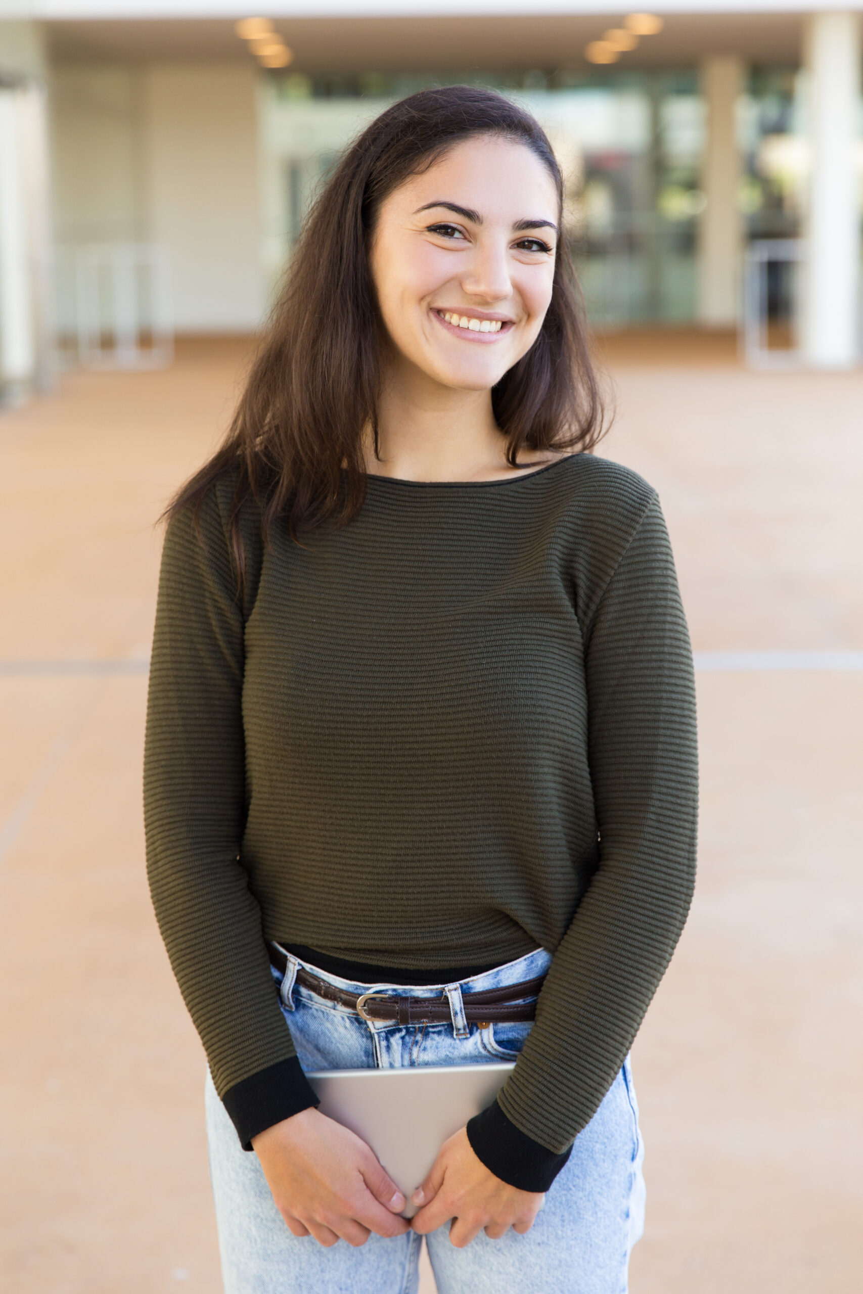 Happy joyful female student with tablet posing indoors. Beautiful young woman in casual standing in hallway, holding digital device, looking at camera, smiling. Gadget user concept